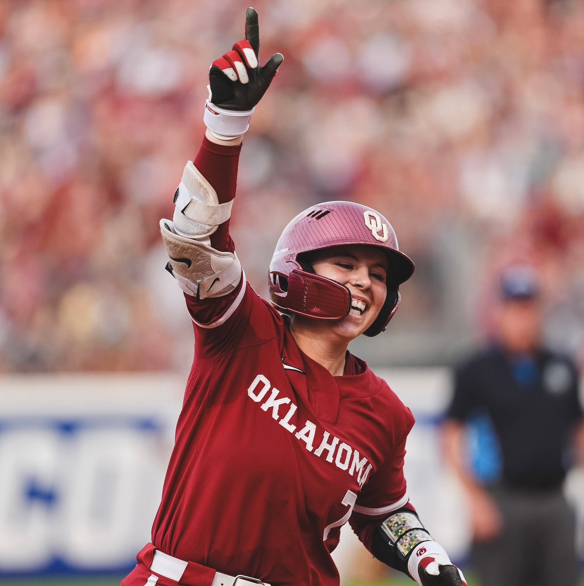 Baseball player in a red Oklahoma uniform celebrating with a raised arm in a stadium setting.
